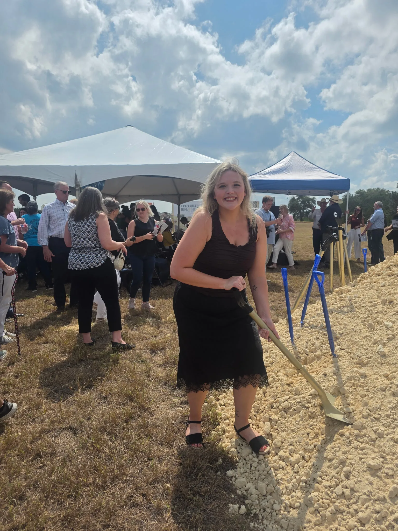 Grace Lakey holding a shovel at a groundbreaking ceremony with white tents and a crowd behind her